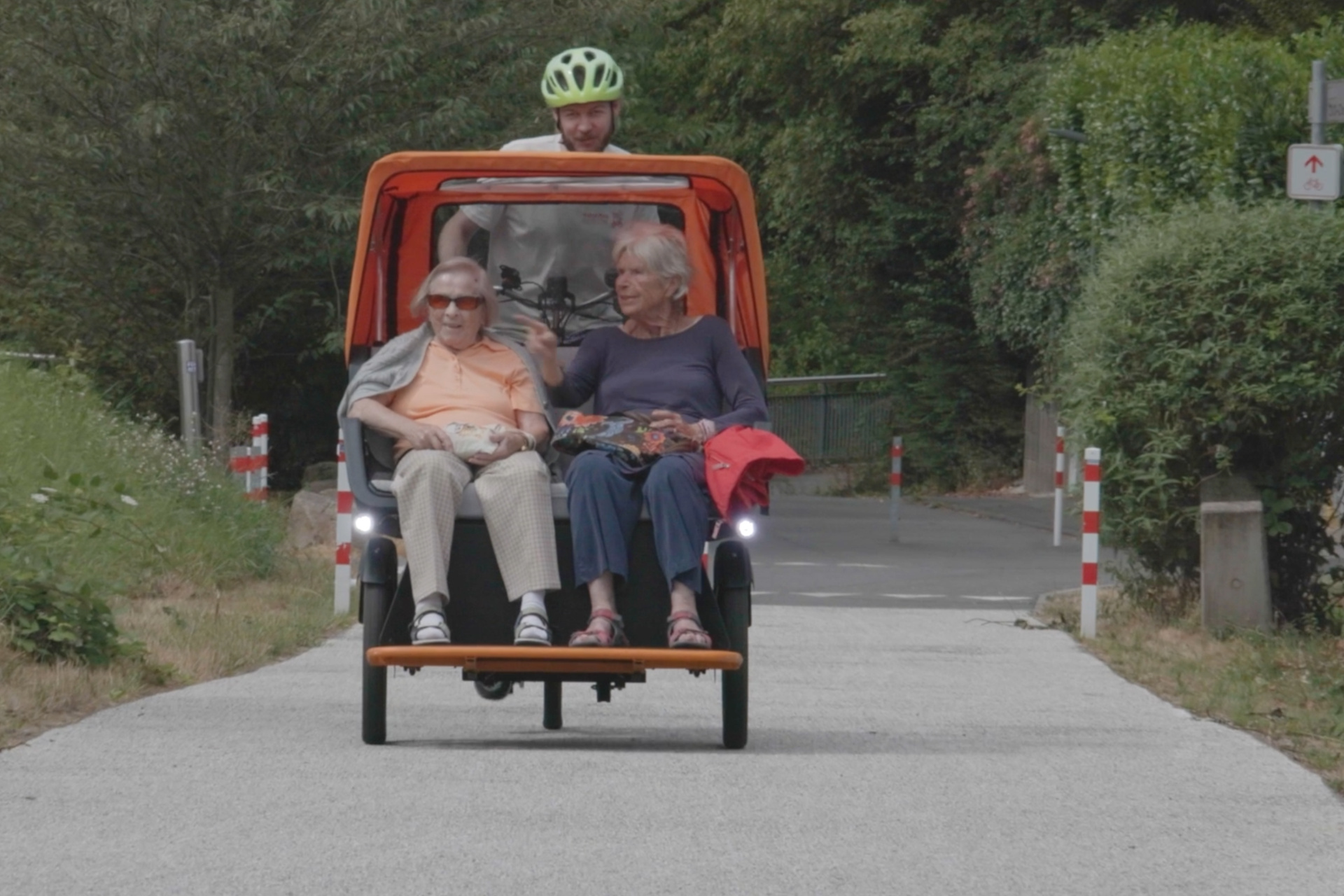 Zwei ältere Damen sitzen vorne auf der Sitzbank einer Rikscha und werden von einem jungen Mann eine leichte Steigung im Grünen hinauf gefahren. Alle wirken entspannt und scheinen die Fahrt zu genießen.